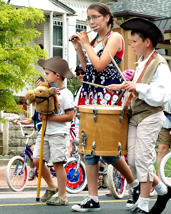 Children marching in a parade dressed in patriotic clothes and playing instruments.